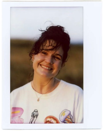 Headshot of Artist: Danika Larade smiles at the camera head tilted and brown bangs wind-blown across their forehead. Danika wears a white t-shirt with painted drawings on it. The background is an out-of-focus grassy landscape with a white grey sky.