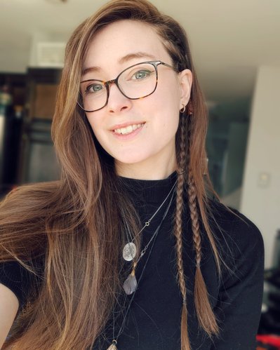 Team Member Headshot: Cassie Seaboyer smiles at the camera, she has two thin braids behind one ear and the rest of her light brown hair frames the other side of her face. She is wearing a black t-shirt and three silver necklaces with pendants.