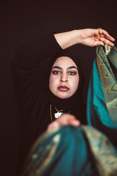 Team Member Headshot: Masuma Khan looks into the camera wearing a black hijab that blends in with her shirt and backdrop. Masuma holds a blue/green and gold designed scarf between her hands; one above her head, the other stretched towards the camera.