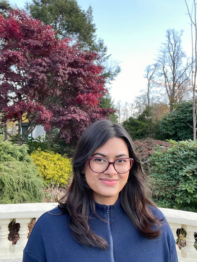Artist Headshot. Rebecca Fernandes smiles at the camera- her brown hair frames her face, she is wearing glasses. In the background is a white stone railing and a garden of bushy hedges and trees. A red maple spreads across the top left half of the  photo.
