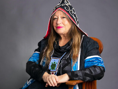 Team Member Headshot: Catherine Martin smiles at the camera at an angle, she wears a black, white, and red pointed hood with intricate detailing, her wavy brown hair hangs down her chest. She sits on a wooden chair against a grey backdrop. 