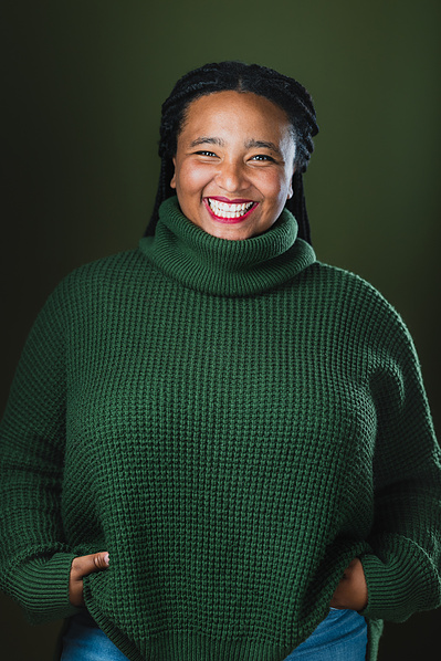 Team Member Headshot: Koumbie smiles at the camera with their hands on their hips in a casual stance. Her black braids are tied back from her face. She wears a green turtle neck sweater and the backdrop behind them is a deep green too.  