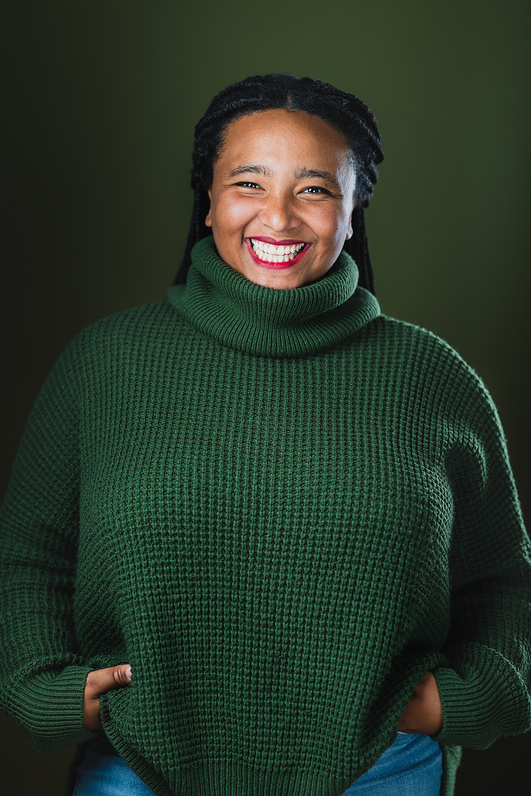 Team Member Headshot: Koumbie smiles at the camera with their hands on their hips in a casual stance. Her braids are tied back from her face. She wears a green turtle neck sweater and the backdrop behind them is a deep green too. 