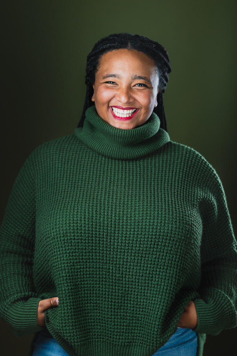 Team Member Headshot: Koumbie smiles at the camera with their hands on their hips in a casual stance. Her braids are tied back from her face. She wears a green turtle neck sweater and the backdrop behind them is a deep green too. 