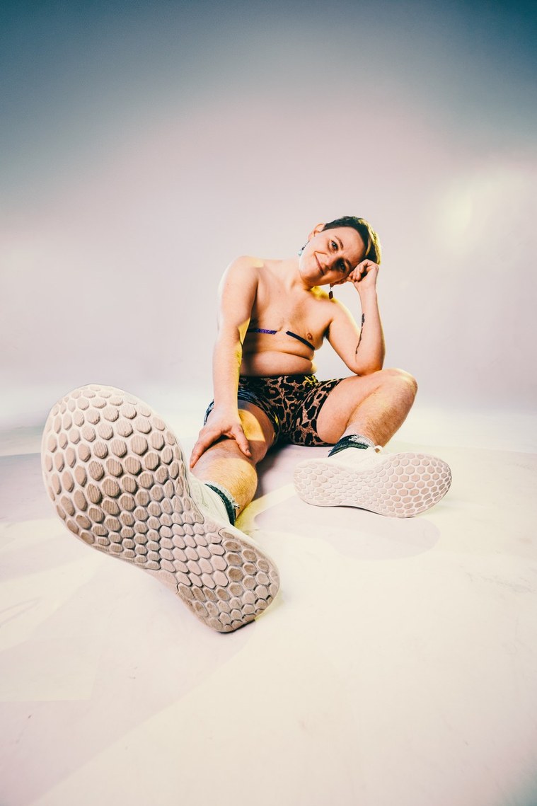 Team Member headshot: Rooks sits on a white backdrop, his pale skin shimmers in the radiant lighting. Rooks’ head is tilted to the side resting on his hand. He has purple glitter tape going across the scars on his chest and is wearing leopard print shorts