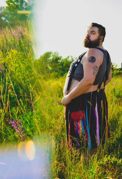 Team Member Headshot: Matthew Downey is standing in a grassy field looking sidelong at the camera.  Matt's brown beard is lined up with their hair. He is wearing a multicolour tassel skirt that is held up with suspenders over a grey crop top. 