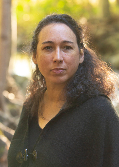 Team Member Headshot: Shalan Joudry looks directly into the camera. Her brown curly hair is pinned back and hangs along her shoulders. Shalan wears a brown sweater. She is standing in front of an outdoor landscape with trees, greenery and water.