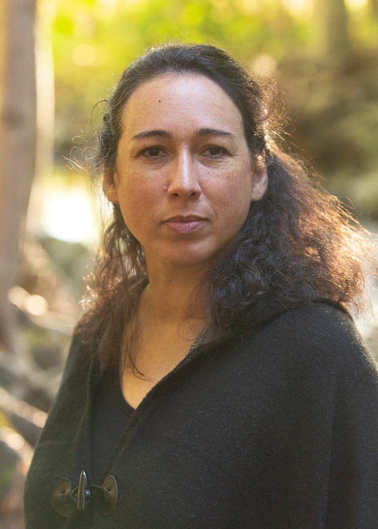 Headshot: Shalan Joudry looks directly into the camera. Her brown curly hair is pinned back from her face and hangs along her shoulders. Shalan wears a brown sweater. She is standing in front of a golden-hour landscape with trees, greenery and water.