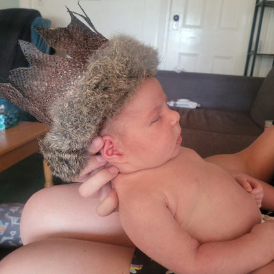 A side-view of baby Nora wearing the finished fish leather crown with rabbit trimmed fur. Nora is sleeping, the crown fits her head perfectly. Her head is being propped up by the hand of the person that holds her.