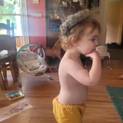 Jane, a young child, wears the fish leather crown with rabbit trimmed fur. Jane is also wearing a pair of yellow pants. The hat rests on the crown of Jane's head. The backdrop is a played-in living room.