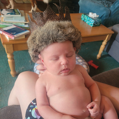 A front-view of baby Nora wearing the finished fish leather crown with rabbit trimmed fur. Nora is sleeping, the crown fits her head perfectly, she wears a diaper. Her head is being propped up by the hand of the person that holds her.