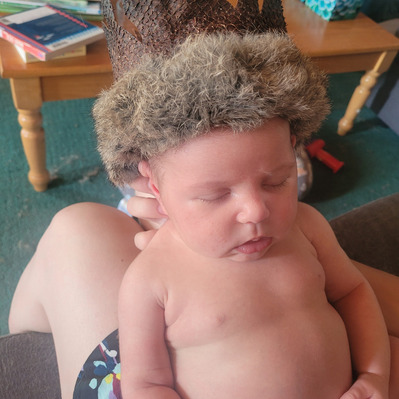A close-up front-view of baby Nora wearing the finished fish leather crown with rabbit trimmed fur. Nora is sleeping, the crown fits her head perfectly. The backdrop is a living room table and the legs of the person holding her.