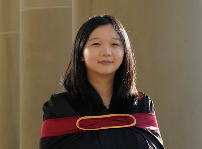 Headshot of Artist: Frisia 李昕怿 looks at the camera with brown-black hair framing her face. Frisia is wearing a black robe with a red band across her chest with a golden oval holding the red band together. The background is brown-beige rolling stone wall. 