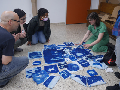 Gallery Image: Many prints are laid on a white dotted floor. The blue of the prints is a vibrant contrast to the rest of the image.  Donica sits on the floor behind the grid explaining something to three onlookers.