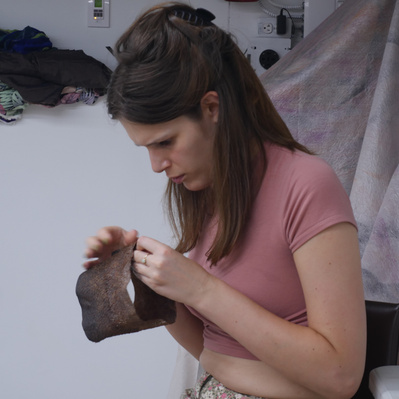 Lara is pictured working on her crown. She is in the process of securing the leather in a cylindrical shape.  The background is a white wall with a folded pile of materials on a shelf behind the artist.