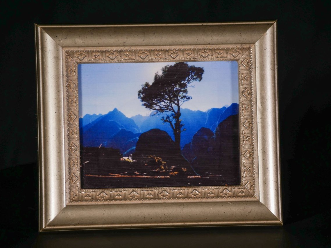 Image of a framed photo on a black background. The frame is gold with angular detailing and sits slightly crooked within the image. The photo is a tall tree cutting into a blue mountain horizon with a stack of wooden logs in shadow along the bottom.