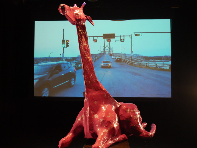 Image is of a papier-mâché model of a Giraffe that sits with its neck & head straight up. It is painted peach w gold stars & moon detailing.  It sits on a shiny black surface and behind them is a projected image of a roadway with street lights. 