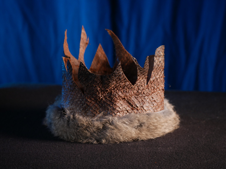 A Salmon leather crown with rabbit fur trim on a black fleece surface in front of a blue curtain. The crown has many points of distinct shapes and sizes. The leather is light brown with a grid-like scaled texture. The rabbit fur is light grey and fluffy.