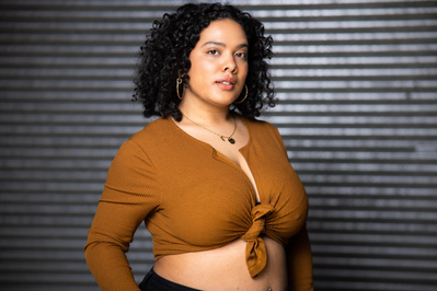 Artist Headshot. Riel Reddick-Stevens looks at the camera at an angle. She wears a brown long-sleeve halter-top that ties together in the centre. Riel's black curly hair frames her face and she wears hoop earrings. Background is a ridged metal wall.  