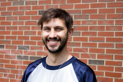 Team Member Headshot: Diego Cavedon Dias smiles at the camera. They wear a white baseball tee, a thin chain necklace and two hooped earrings. Behind Diego is a brick wall. His cropped brown hair has a side part and he has a short brown beard.