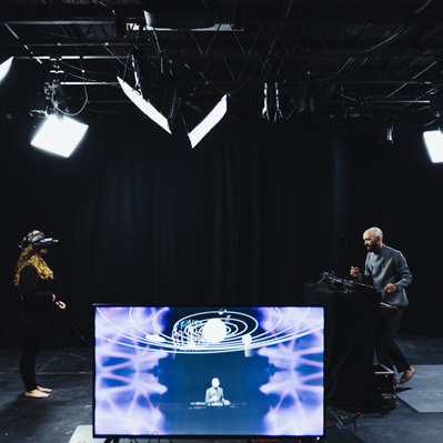 In the background Sylvia Bell stands on the left wearing a VR headset looking across the room at Aaron Collier who is standing in front of synthesizers. In the foreground  a TV screen showing Aaron augmented with a VR solar system and psychedelic textures
