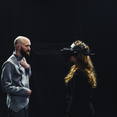 Aaron Collier and Sylvia Bell stand facing each other backgrounded by black. Aaron on the left, bald and wearing a grey suit, his hands on his chest and stomach. Sylvia on the right, long wavy hair, wearing a VR headset.