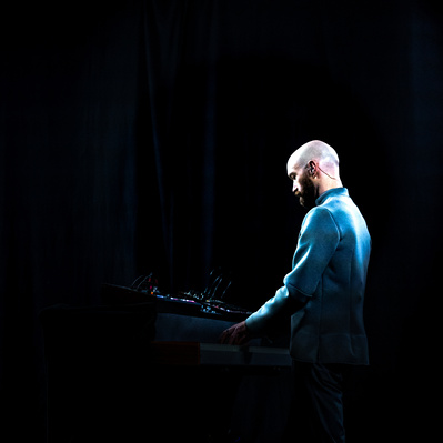 Aaron Collier is wearing a grey suit and is picked out of a black background with a light somewhere above him. He is turned to the side and slightly away from the camera and is playing his keyboards.