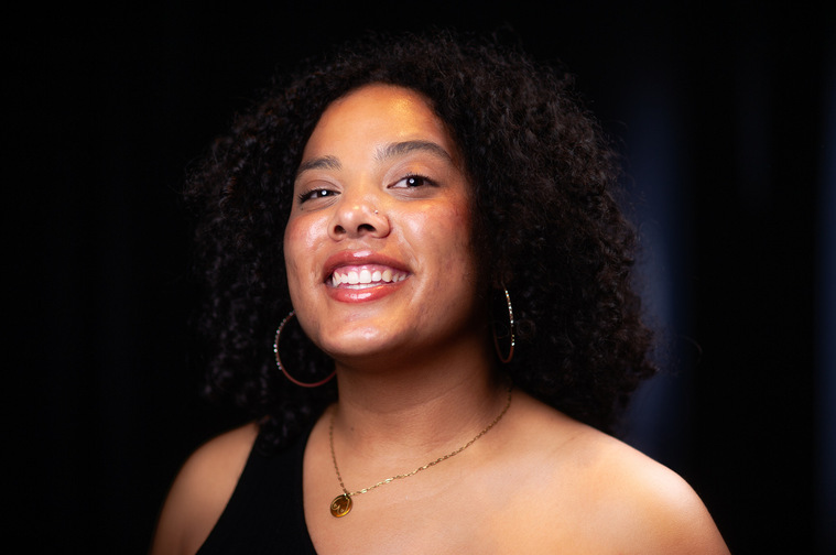 Artist Headshot. Riel Reddick-Stevens looks at the camera at a slight angle. She wears a black top with one shoulder strap and a pendant on a thin chain necklace. Riel's black curly hair frames her face and the background is black curtains.