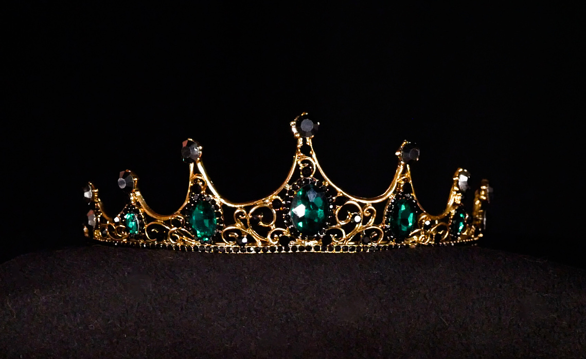 Image of an ornate golden crown on a black background. The crown has seven points with the centre point being the highest. Each point has a black circular gem encrusted within it.  Under each point is a green gem. The crown glistens under lights. 