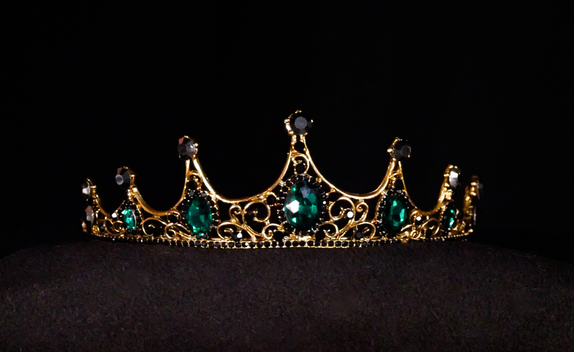 Image of an ornate golden crown on a black background. The crown has seven points with the centre point being the highest. Each point has a black circular gem encrusted within it.  Under each point is a green gem. The crown glistens under lights. 