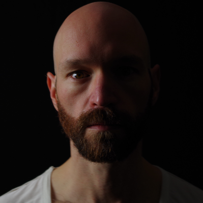 Aaron Collier looking directly at the camera with a serious face. He is bald, has a beard, is wearing a white t-shirt and is light dramatically from his right side.