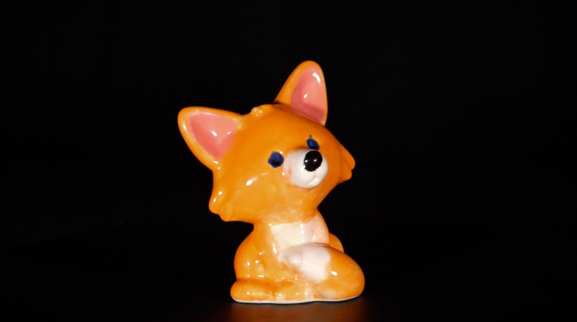 Image of a small porcelain fox figurine on a black background. The fox is cartoonish and sits up like a human on bent knees. The fox has black painted eyes and nose on white muzzle. The belly of the fox is also painted white along with the tip of its tail