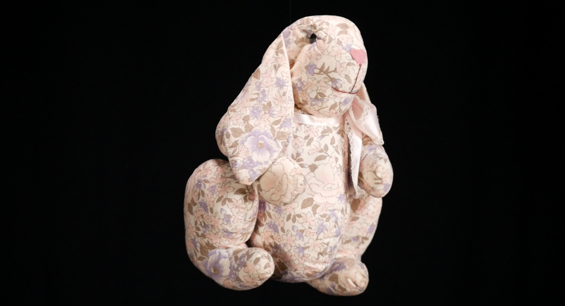 Image of an ornate stuffed bunny on a black background. The children's toy looks hand-sewn with a light pink floral printed material. The face of the bunny is created with sewn lines. The bunny wears a pink ribbon with lace-fringe around its neck.