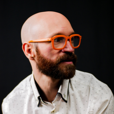 Team Member Headshot: Aaron Collier looks to the side, we see his profile with a red-brown beard and bald head. Aaron wears a white polka dot button up shirt and bright orange glasses. Aaron floats on a black backdrop.