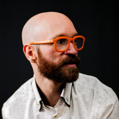 Headshot: Aaron Collier looks to the side, we see his profile with a red-brown beard and bald head. Aaron wears a white polka dot button up shirt and bright orange glasses. Aaron floats on a black backdrop.
