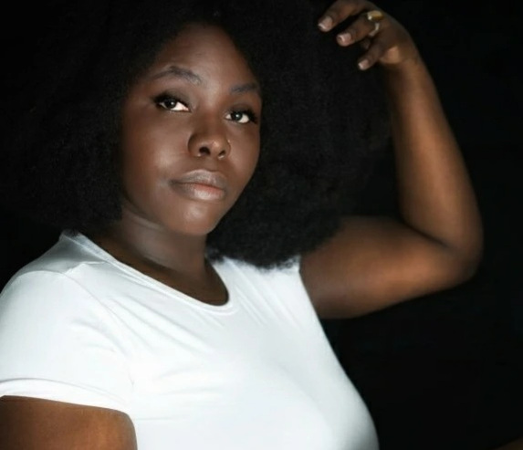 Artist Headshot. Nailah looks at the camera with her hand above her head touching the top of their hair. She wears a white t-shirt. Nailah's black curly hair frames their face on a black background.