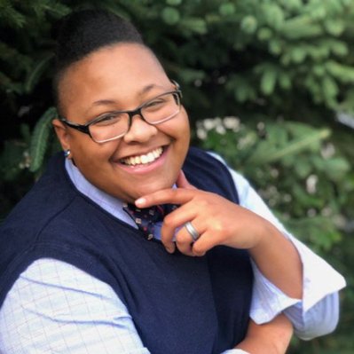 Team Member Headshot: Evelyn Bradley smiles at the camera with her hand on her chin in an L shape. Evelyn wears a blue sweater vest over a white grid-lined dress shirt with a patterned bow-tie.  Her hair is in a crisp pompadour shape.