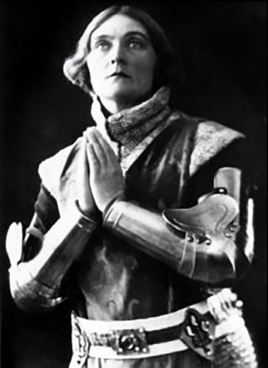 Black and white image of Sybil Thorndike with her hands together in a prayer position. She wears armour with arm plates, winged shoulder plates and a belt with many cotes of arms. Sybil looks above the camera her face half in light, half in shadow.