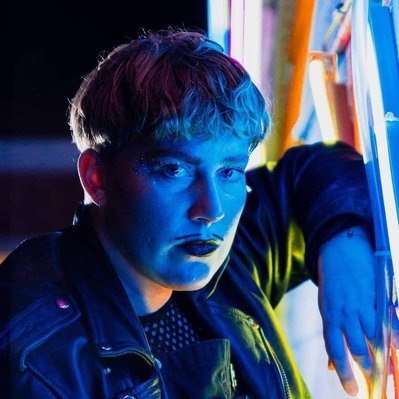 Headshot of Artist Lou Campbell. Lou rests with their arm up against a brightly lit wall with lots of neon lights. They look into the camera at an angle. Lou's face is bathed in blue & orange light. Lou has a dark lip colour and a drawn on moustache.