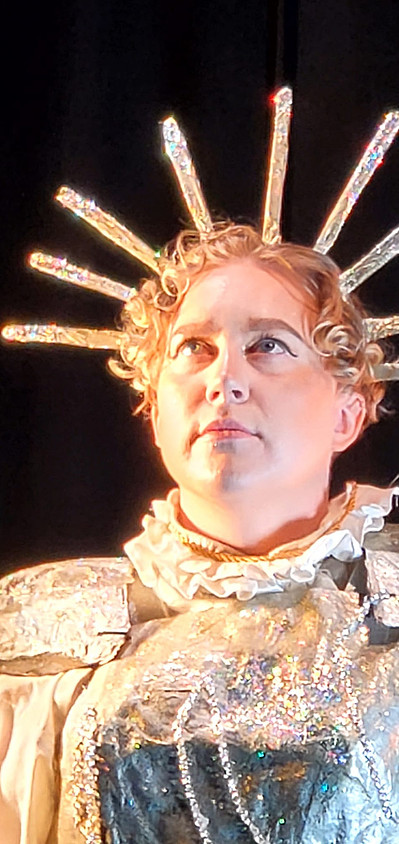 Headshot of Lou Campbell in their crown, ruffled collar and chest plate. They look up to the left corner of the image with a solemn expression. Lou's strawberry blond ringlets frame their forehead. The light colours in the image are particularly vibrant. 