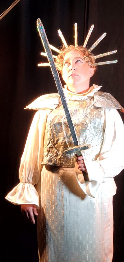 Lou Campbell wears a crown with nine foot-long points radiating outwards in the shape of a rainbow. They hold a sword out in front of them. Their outfit includes a ruffled collar, shoulder plates and a chest plate. Under the armour is a white lace robe. 