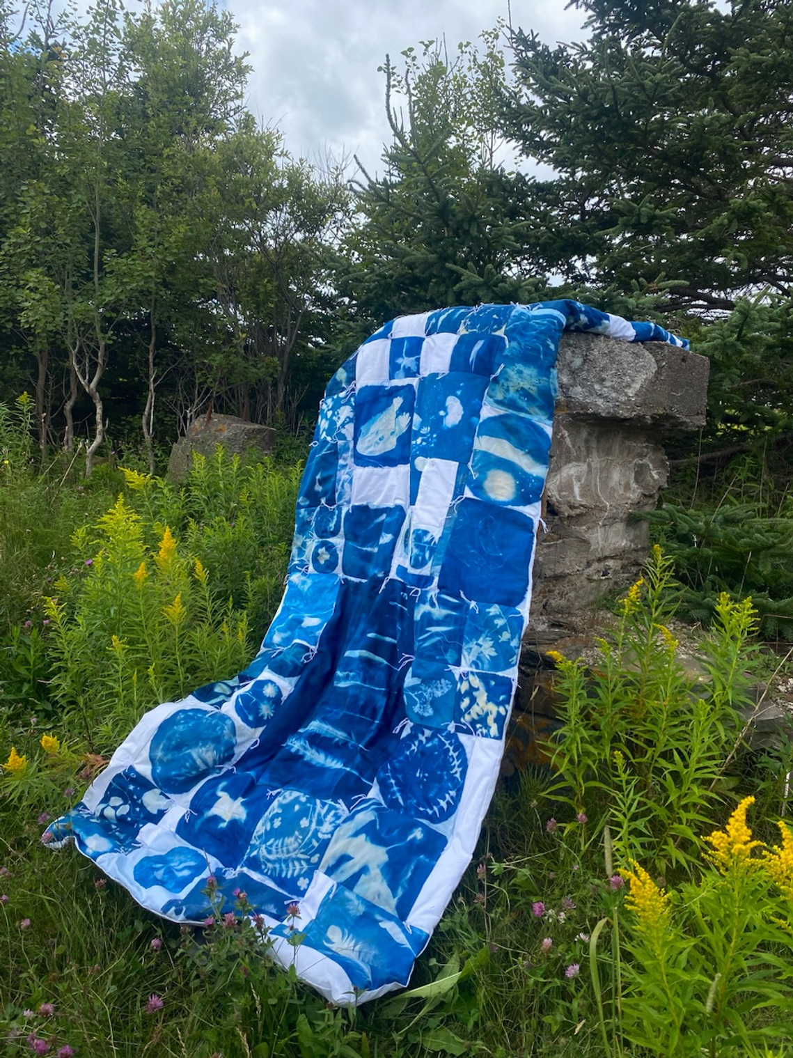 Donica's quilt, made of cyanotype prints, is draped over the ruin of an old stone fence. Forest greenery and goldenrod surround the quilt as its blue fabric shows up in vibrant contrast. There are many patches on the quilt with various prints.