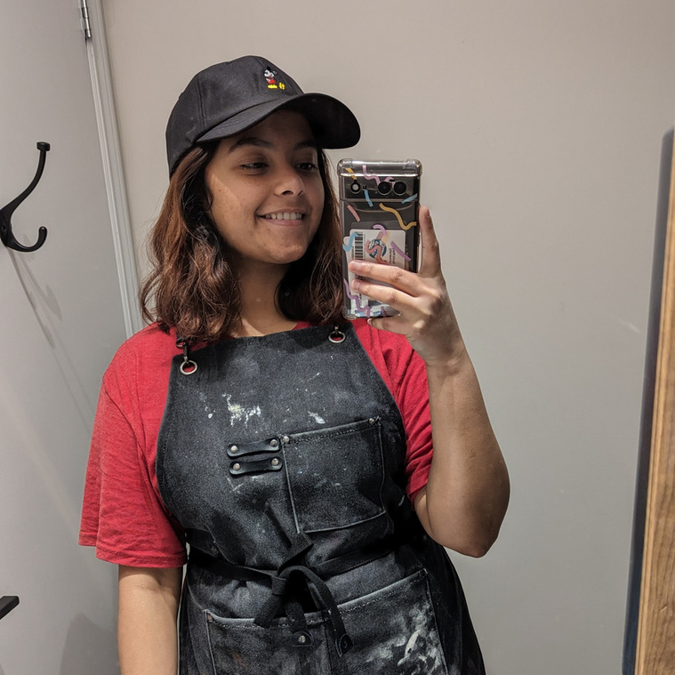 Artist Headshot. Urvi Bhatt wears a black artist's smock and a black ball cap in front of a mirror. Urvi smiles at the camera and holds up her phone with her left hand. The mirror is in a light brown frame and the rest of the room is painted white. 