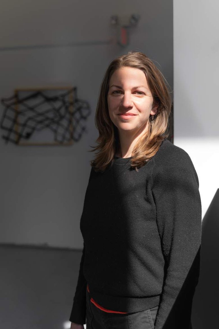 Portrait of artist Catherine Nelson by photographer Rafael Concepción.  She stands in the foreground of her exhibition with shoulder-length brown hair and sunlight cast across half of her body next to a sculpture shaped like a fishing net.  