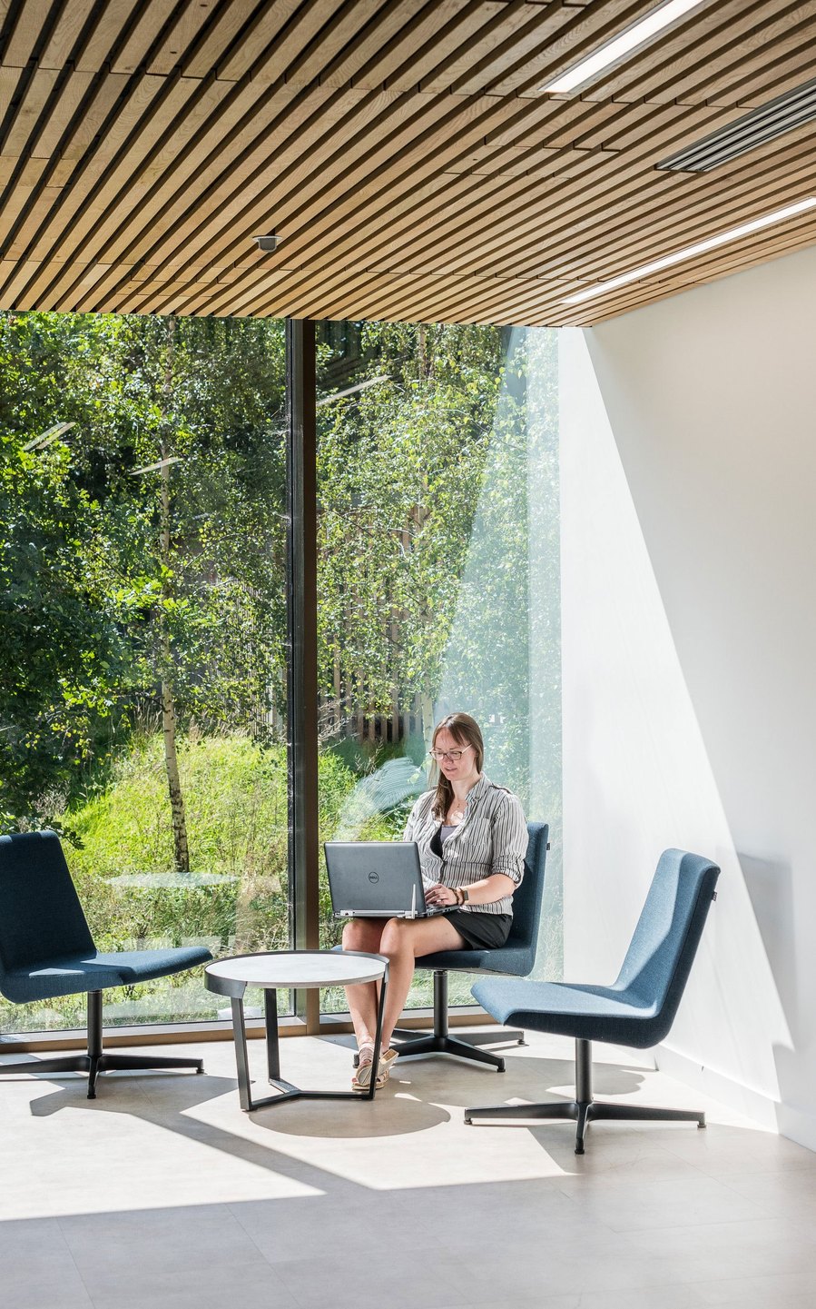 Person sitting at a table with a laptop in a modern room with large windows overlooking greenery.
