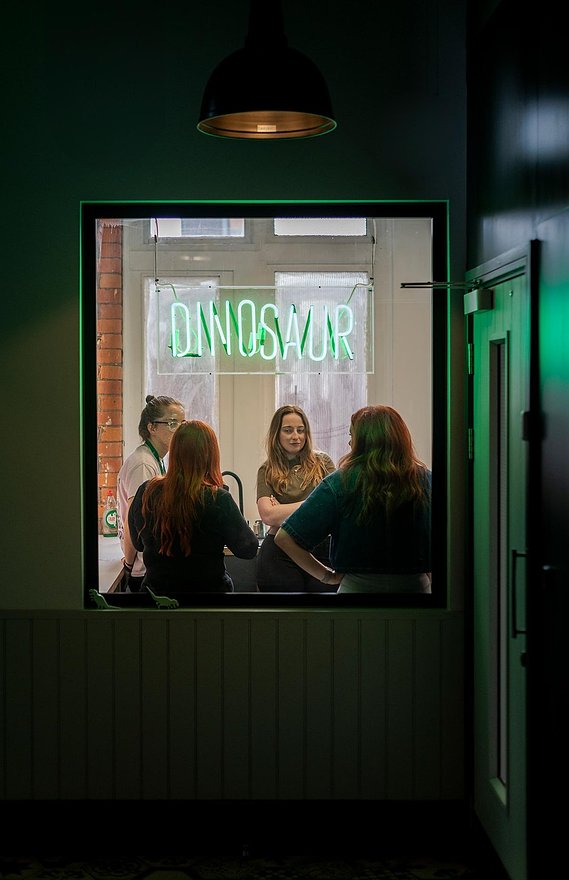 commercial lifestyle photography showing staff in a workplace, candid photograph shot through window, team members stood in kitchen area, neon sign in window 