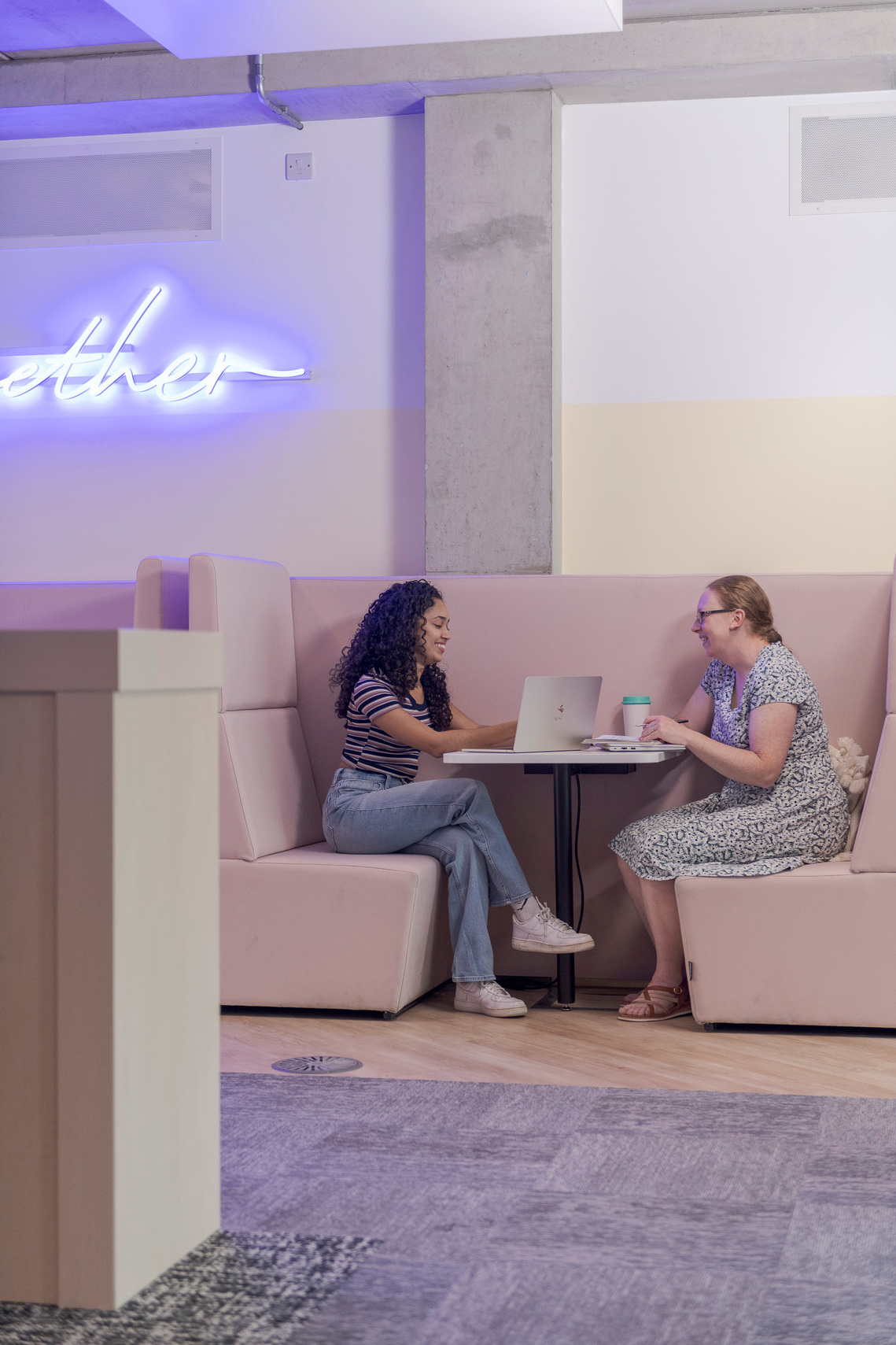 a more informal education lifestyle photograph, with one student and an academic sat discussing work. neon sign on wall hints to modern interior design