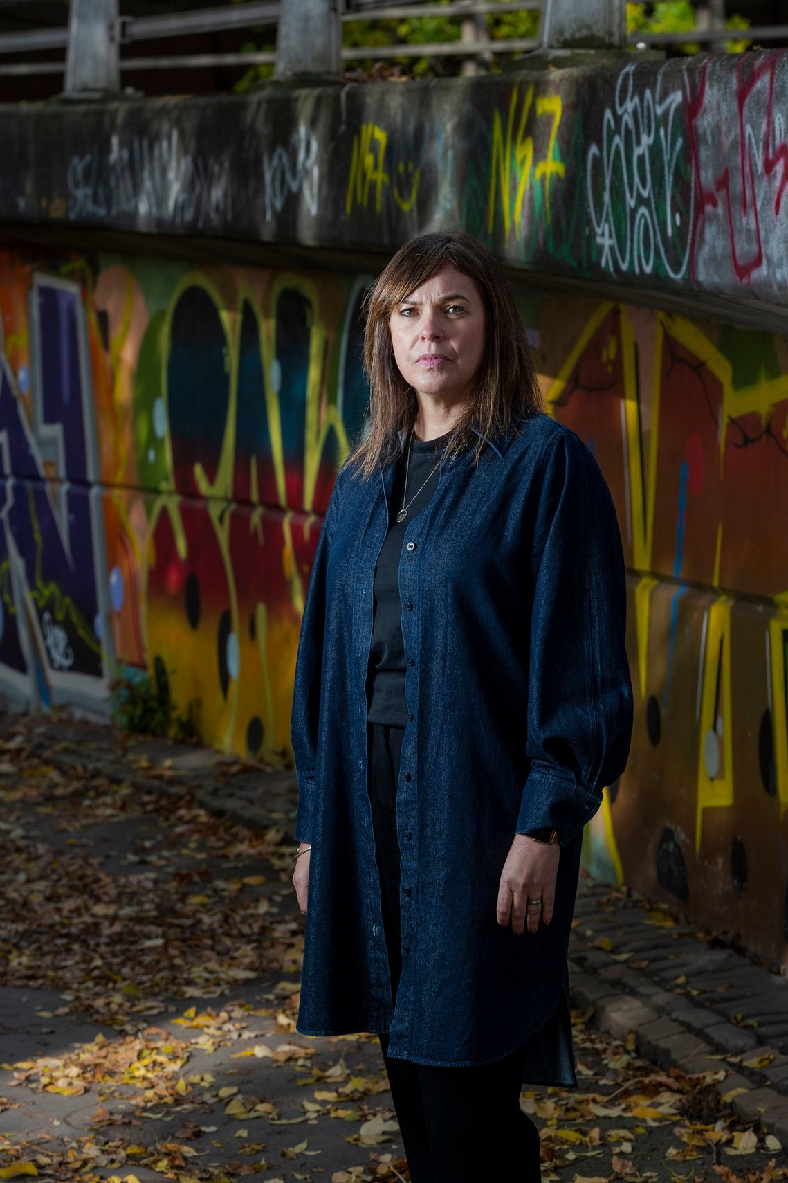 A woman stands in front of graffiti. She wears a dark denim shirt and black top, appearing thoughtful. Captured by a Manchester freelance photographer.