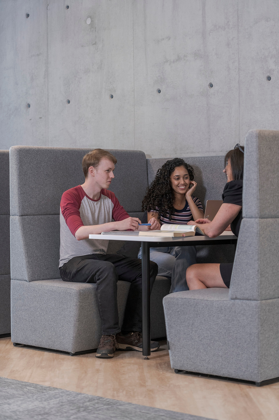 two students and tutor sat in a booth with books and laptops, in discussion about a project and smiling