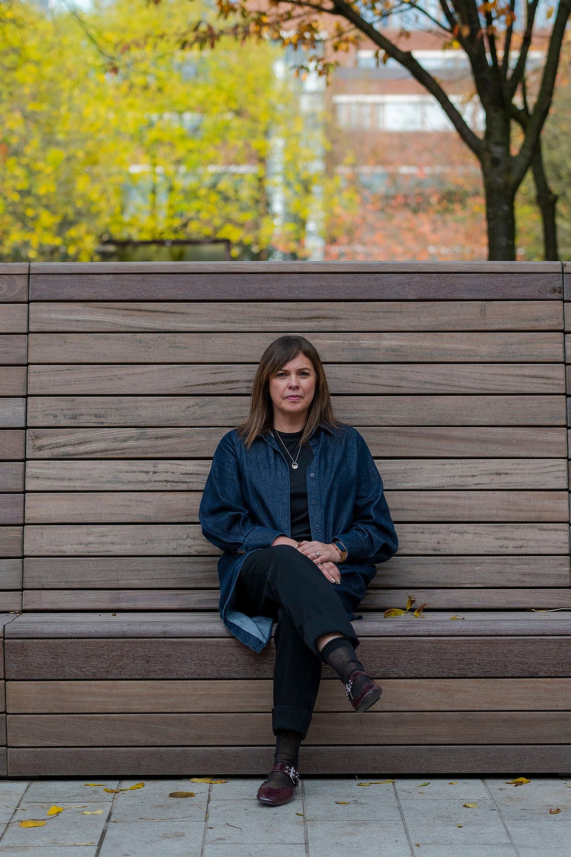 A woman sits on a large wooden bench in a park, surrounded by autumn foliage.
